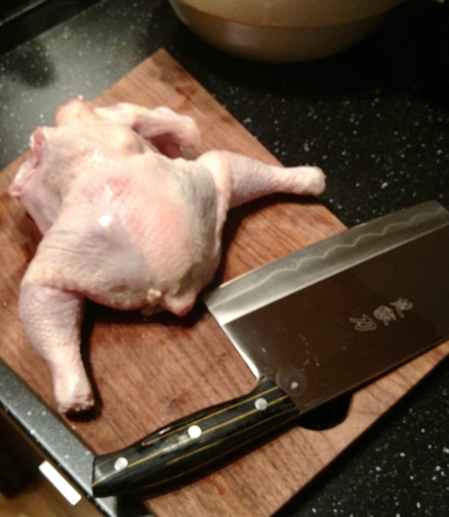 Raw whole chicken on top of a cutting board next to a Chinese cleaver.