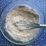 cocoa powder, flour, salt, baking powder, and espresso measured into a glass mixing bowl and whisked to blend the ingredients with the whisk still in the glass mixing bowl sitting on top of a dark blue with multi-colored polka dots