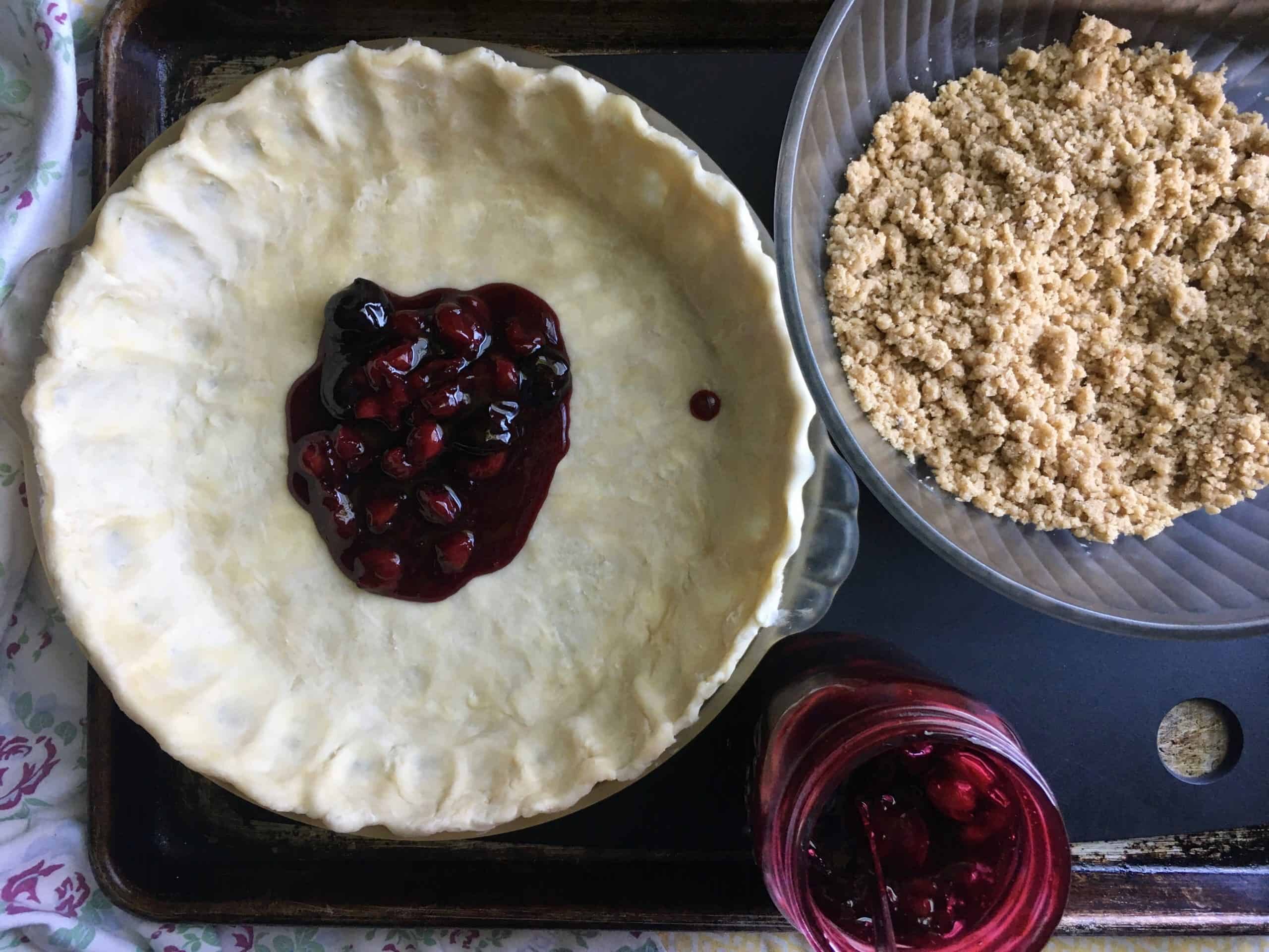 an unbaked pie crust with a ladle full of homemade cherry pie filing in the middle with the crumb streusel topping in a bowl to the right and the mason jar of cherry pie filling in between