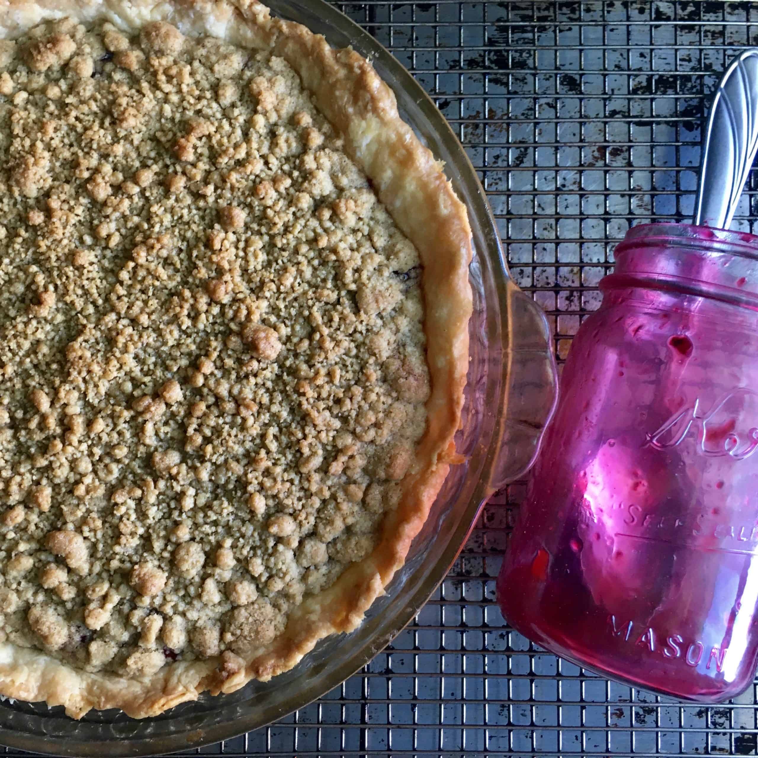 whole cherry crumb crunch pie on a cooling rack with a cherry pink stained mason jar with a large spoon in it lying next to the pie on it's side