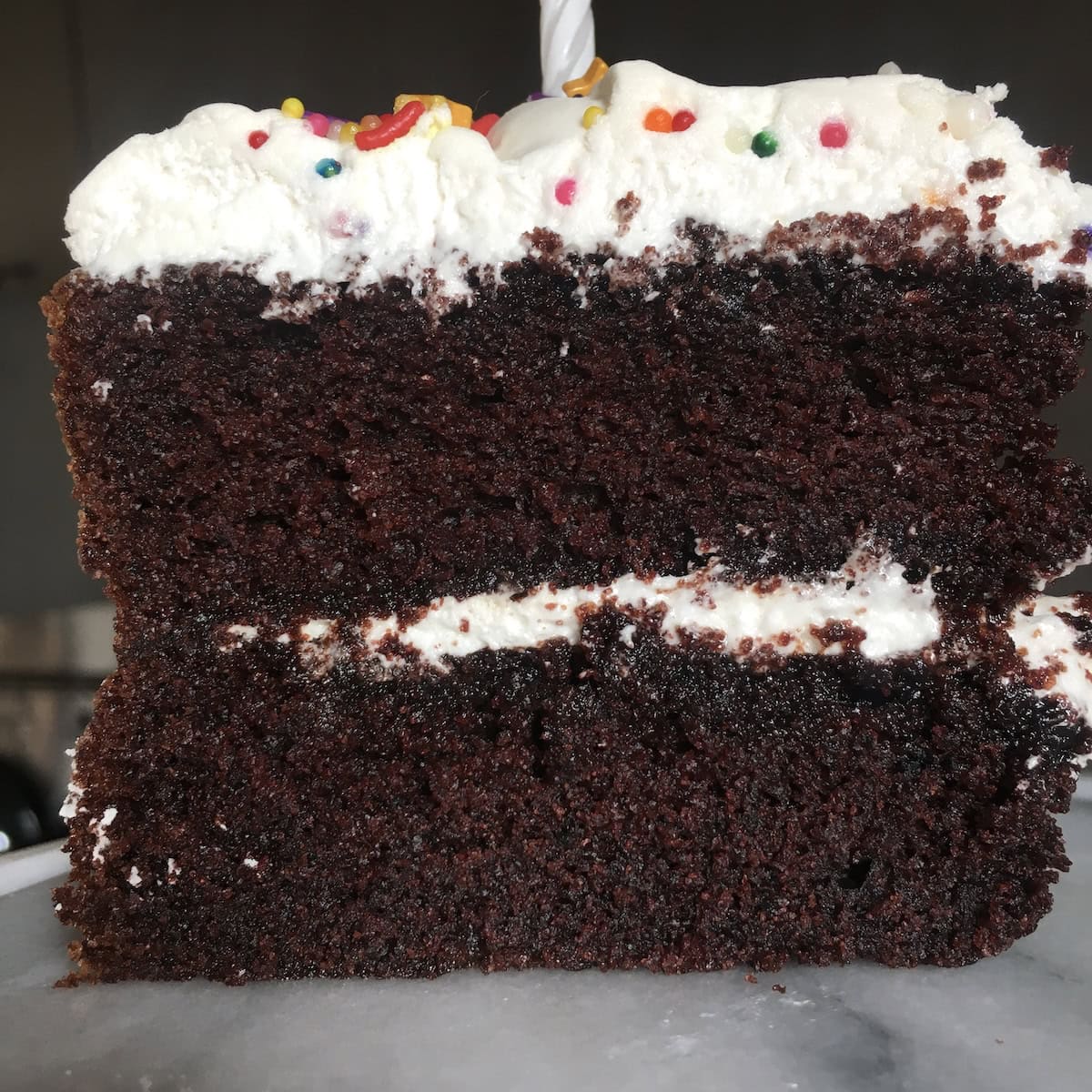 Double layer devil's food cake with 7-minute frosting and birthday candy sprinkles with a lit candle, close up showing the super moist crumb of this best devil's food cake recipe from scratch for chocolate birthday cake.