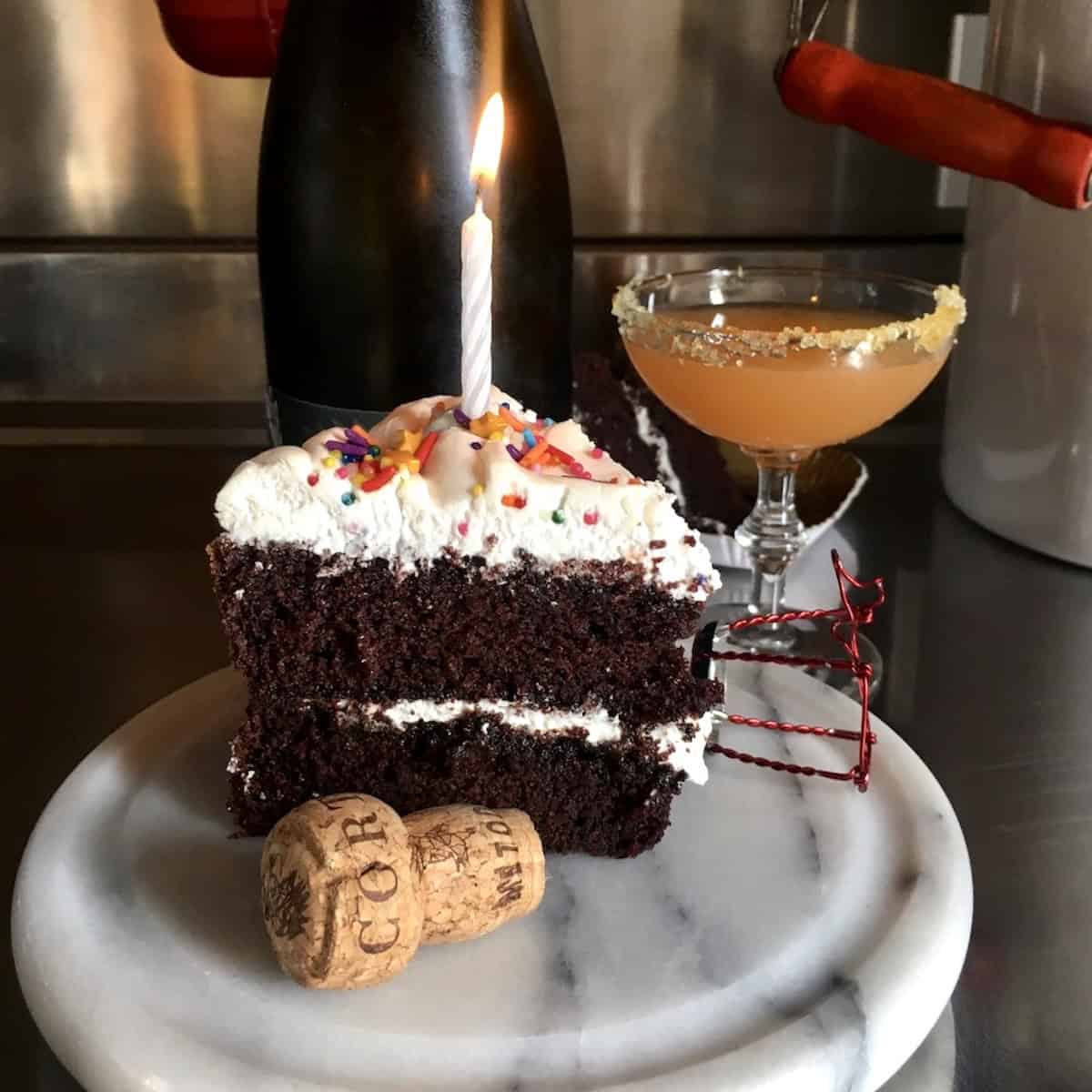 Best devil's food cake slice with 7-minute frosting, birthday sprinkles, and a lit candle on a marble tray next to a homemade champagne cocktail with homemade orange sugar rim, the best chocolate devil cake for birthday celebrations and special occasions.