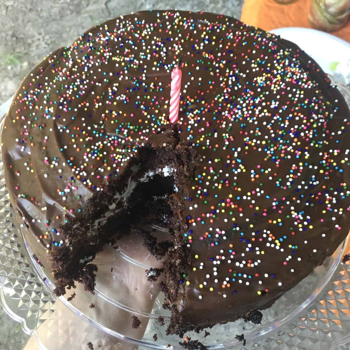 Homemade oversized ding dong cake made with moist devil's food cake, creamy filling, and chocolate ganache topped with nonpareil birthday sprinkles and a lit candle with a slice removed showing the dark chocolatey crumb, the best devil chocolate cake for birthday celebrations.