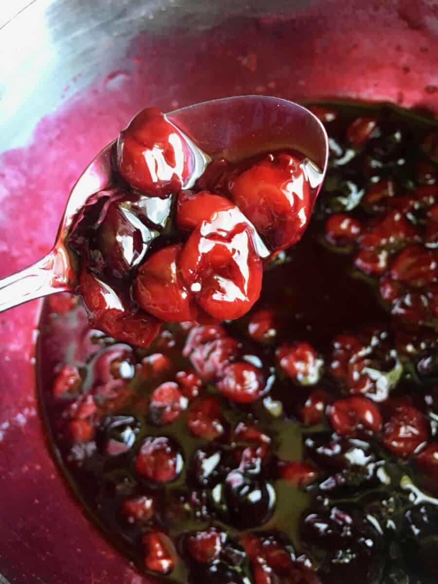 a perfect spoonful of dark and bright red cherry pie filling on a silver serving spoon being held above the pot with the rest of the cherry pie filling below