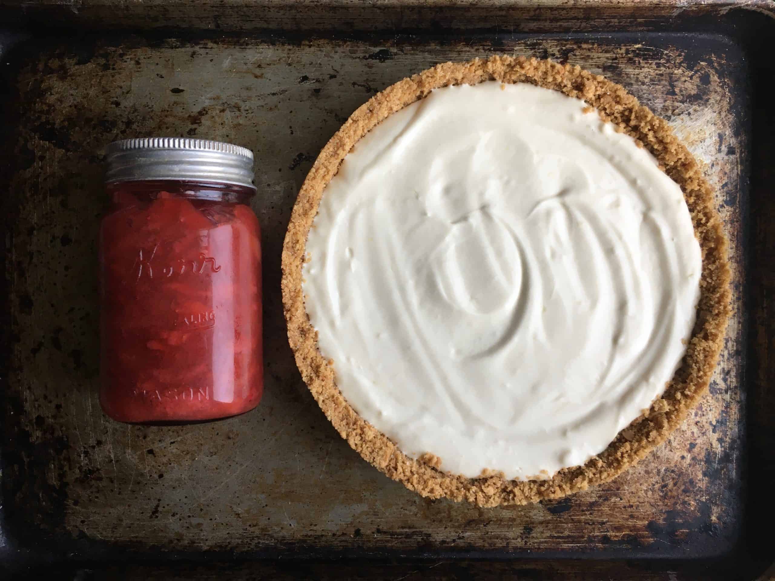 a mason jar filled with homemade strawberry rhubarb pie filling lying to the left of the set up cream cheese pie waiting to be topped