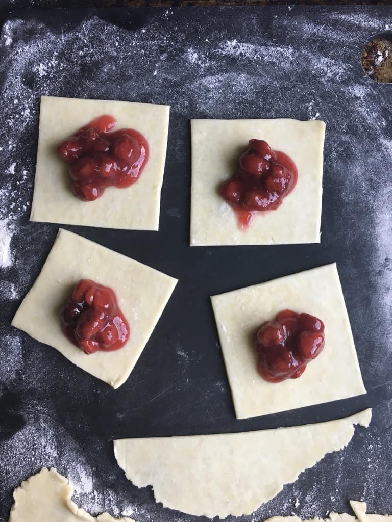 squares of fried pie dough on a black cutting board filled with about 2 tablespoons of cherry pie filling in the middle