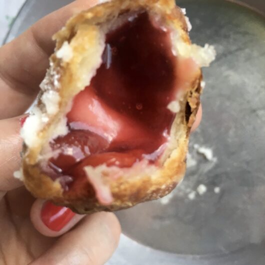 my hand holding a cherry fried pie wonton with a bite taken out and it's gloriously cherry red filling exposed inside