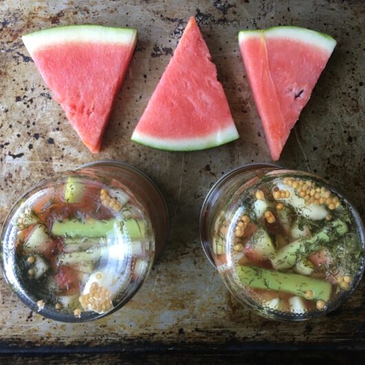 3 wedges of fresh watermelon slices on a sheet pan with two jars of pickled watermelon rinds turned upside down revealing the dill and lemongrass in the jars