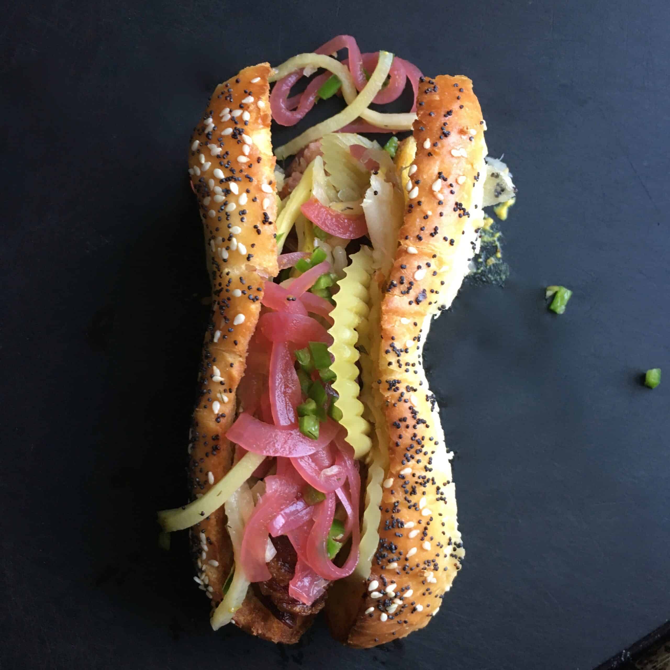 bacon wrapped and shallow friedall beef hot dog with homemade pickled smokey red onions, small diced fresh jalapeno, sauer kraut and dill pickled green tomato slices on a homemade sesame poppyseed hot dog bun