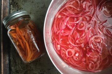 sliced magenta-pink colored red onions in a vinegar based brine inside a stainless steel pot with a sealed Mason jar to the left holding freshly pickled julienned carrot slices