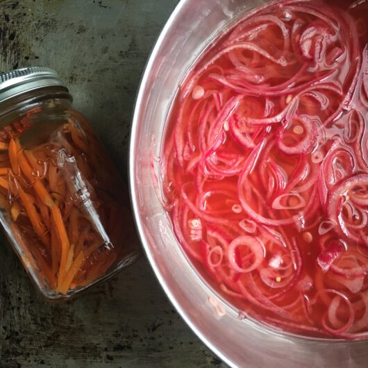 sliced magenta-pink colored red onions in a vinegar based brine inside a stainless steel pot with a sealed Mason jar to the left holding freshly pickled julienned carrot slices