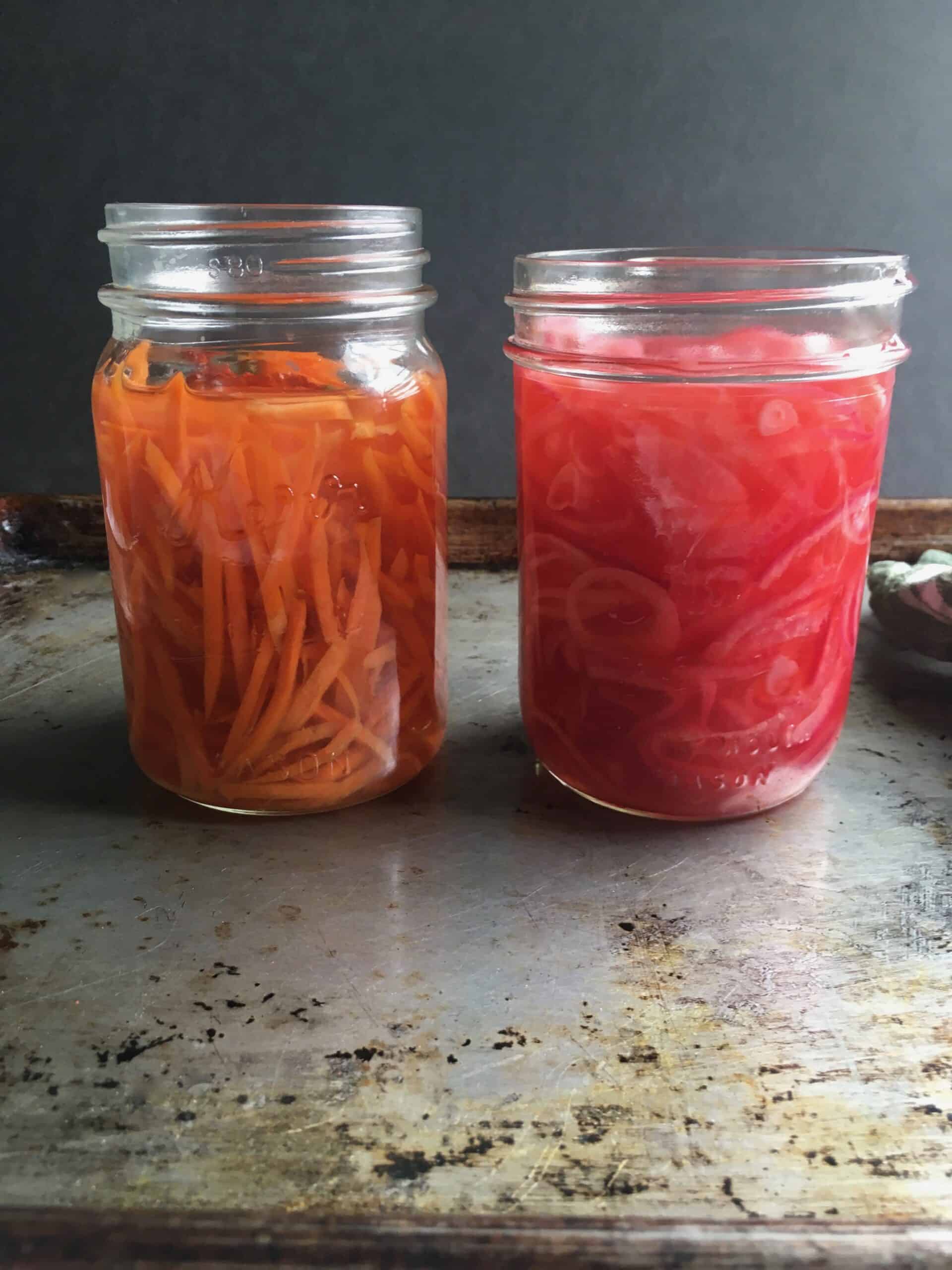 top down view of spicy pickled carrots in a mason jar on the left and a jar of smoky pickled red onions on the right with a green and white checkered tea towel off to the right of the onions