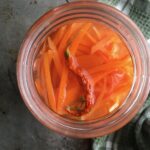 pickled julienned carrot strips in a widemouth Mason jar with a Sichuan bird's eye chili floating on top and a green and white plaid tea towel lying next to it on a sheet pan