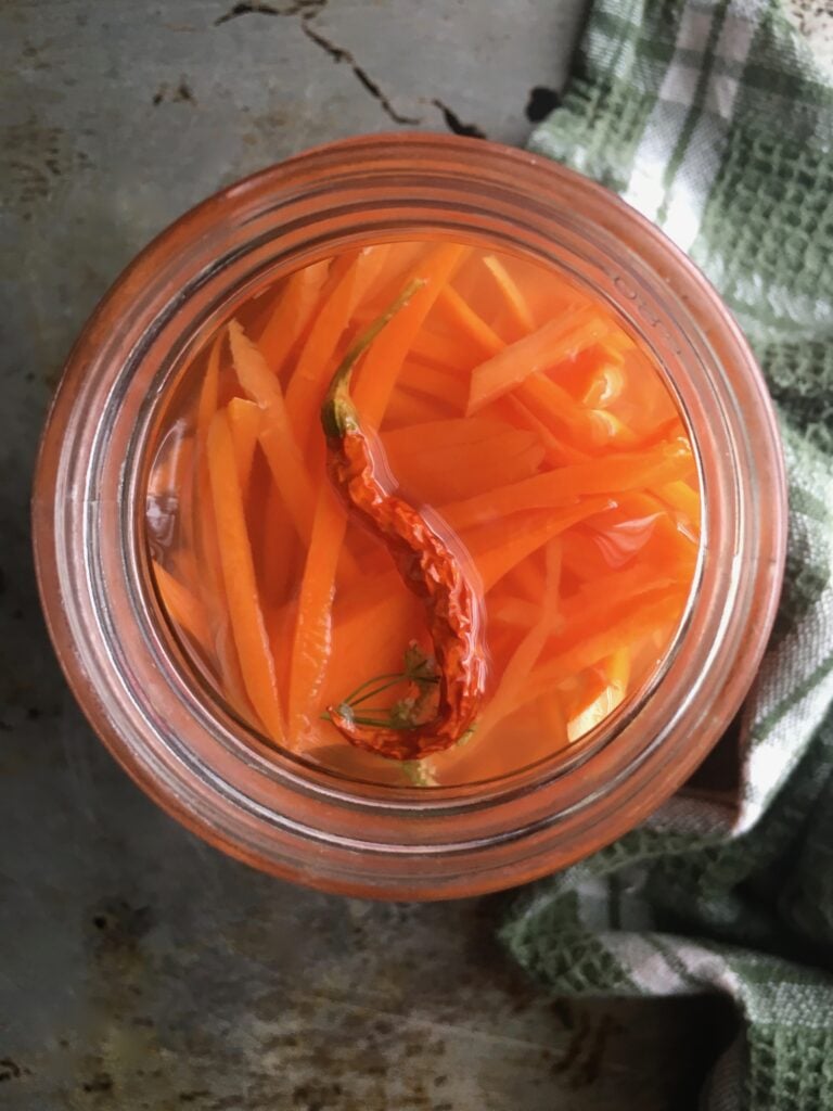 pickled julienned carrot strips in a widemouth Mason jar with a Sichuan bird's eye chili floating on top and a green and white plaid tea towel lying next to it on a sheet pan