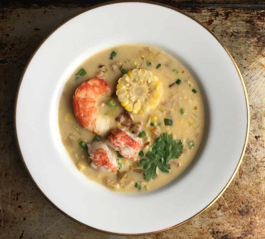 top down view of a bowl of corn chowder with lobster tail, sliced corn on the cob piece with an argentinian royal red shrimp and sprinkling of chives and parsley
