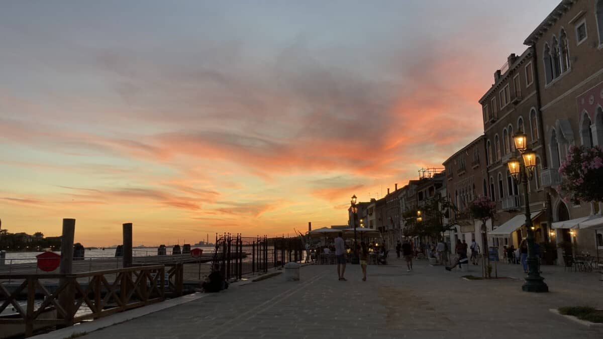 a gold, orange, pink, and turquoise sky in Venice near the water's edge with people dining in the distance as the sun goes down