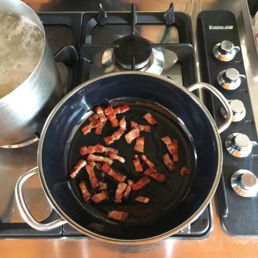 a braising skillet with two handles with lardons of beautifully browned pancetta cooking with pasta boiling in the background