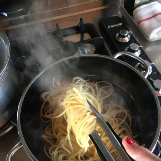 a hand and tongs tossing the noodles in the skillet with the fat and oils