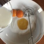 sugar and honey and 2 bright orange egg yolks in a bowl and a measuring cup pouring milk into the bowl