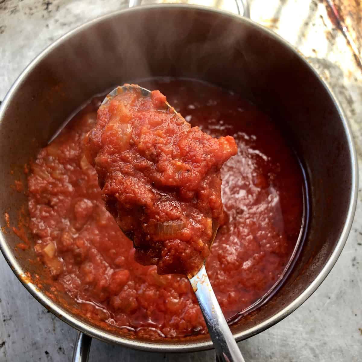 Deep red spicy homemade amatriciana sauce with crispy guanciale on a spoon above the pot, authentic Italian pasta sauce recipe.
