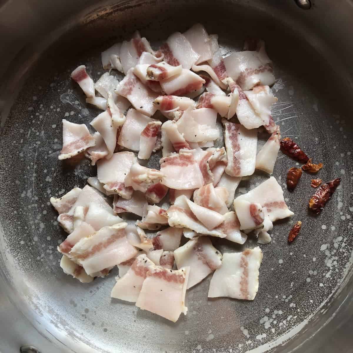 Raw guanciale cured pork jowl with whole red peppers in skillet starting to render fat, how to make amatriciana sauce recipe.
