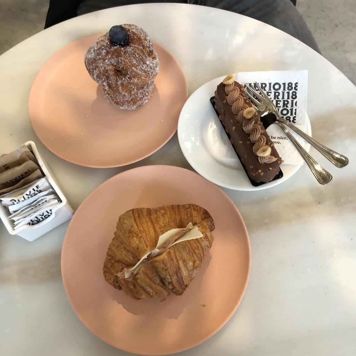 An chocolate-caramel-nougat dessert, a blueberry crossiant-like sweet pastry, and a hot melty savory croissant with prosciutto cotto and provolone on the table with Olivieri 1882 napkins from Olivieri's pastry shop in Arzignano, Italy.