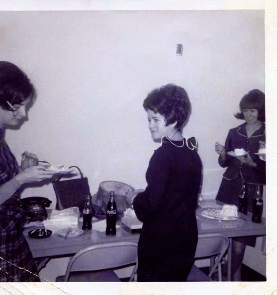 a black and white photo of my Mom wearing a black dress with scalloped collar with white piping standing in front of a table with all kinds of stuff on it with two friends on either side as she's getting a coke or something at her wedding shower
