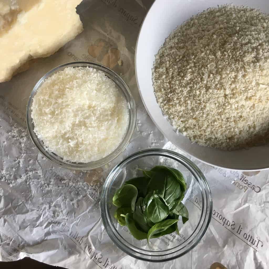 a small bowl of basil leaves and small bowl of grated parm with the huge chunk of cheese in the back and a medium bowl with panko bread crumbs all on top of cheese paper