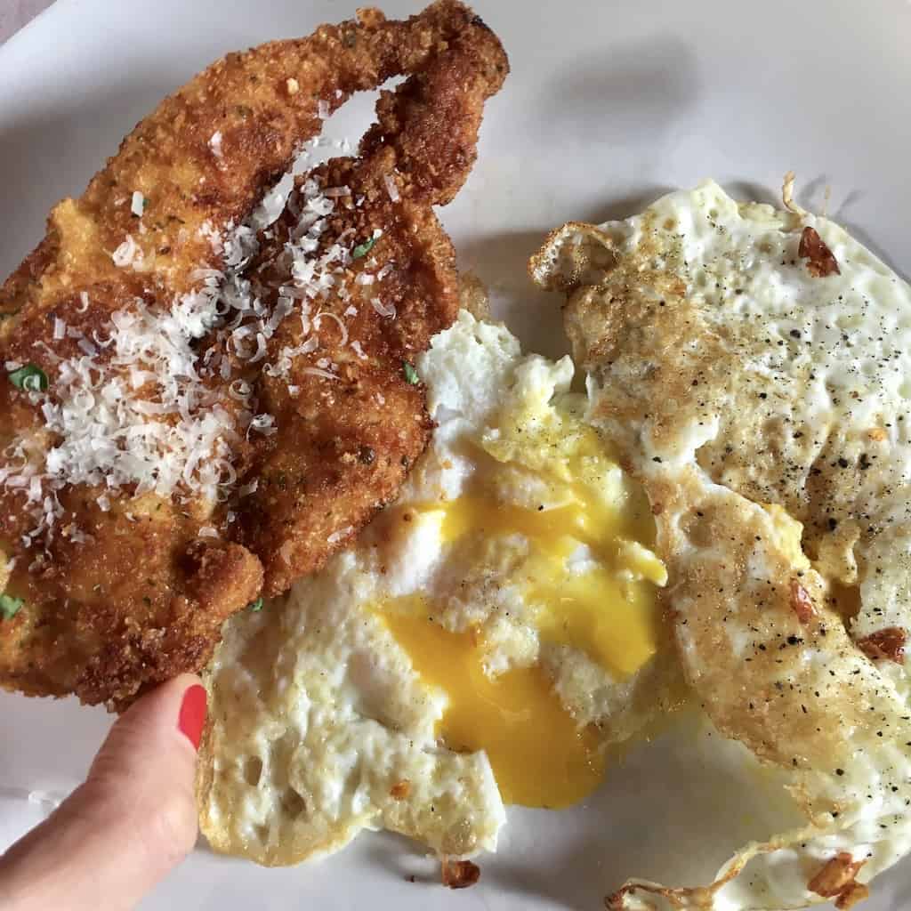 hand holding crispy fried chicken cutlet with fried eggs with golden yolk spilling out in the background