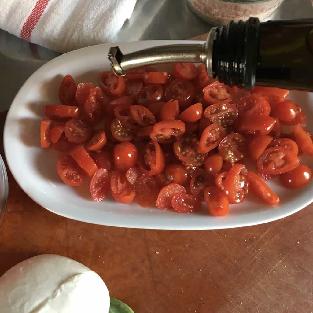 freshly sliced grape tomatoes on a white oval platter getting drizzled with olive oil with a ball of mozzarella in the foreground