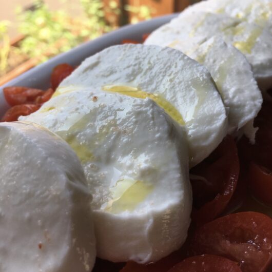 thick milky luscious slices of mozzarella lying on top of a bed of datterino grape tomatoes on a white platter with a drizzle of olive oil and himalayan pink salt over the top with plants in the background