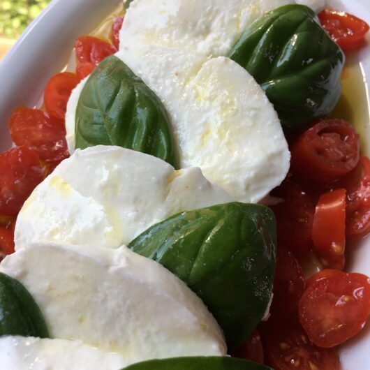a caprese salad on a white oval platter with fresh slices of fresh milky mozzarella, sliced datterino tomatoes, olive oil, fresh basil and sea salt by a window with just a hint of the herb garden on the left side
