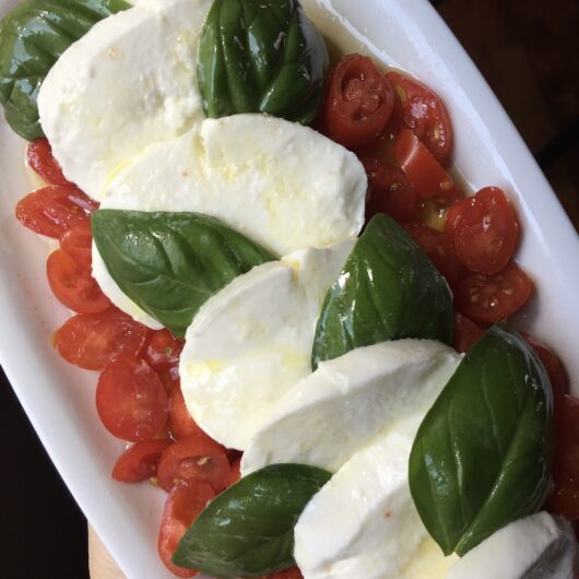 a caprese salad on a white oval platter with fresh slices of fresh milky mozzarella, sliced datterino tomatoes, olive oil, fresh basil and sea salt by a window with just a hint of my hand showing