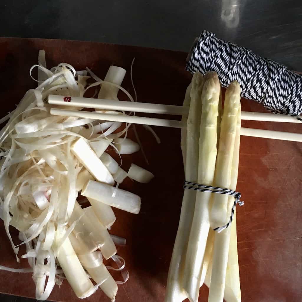 a pile of asparagus skin peels and tough ends lying next to the peeled and bundled asparagus with black and white baker's twine roll above and a pair of Huan Ting chopsticks resting in the middle of the asparagus bundle