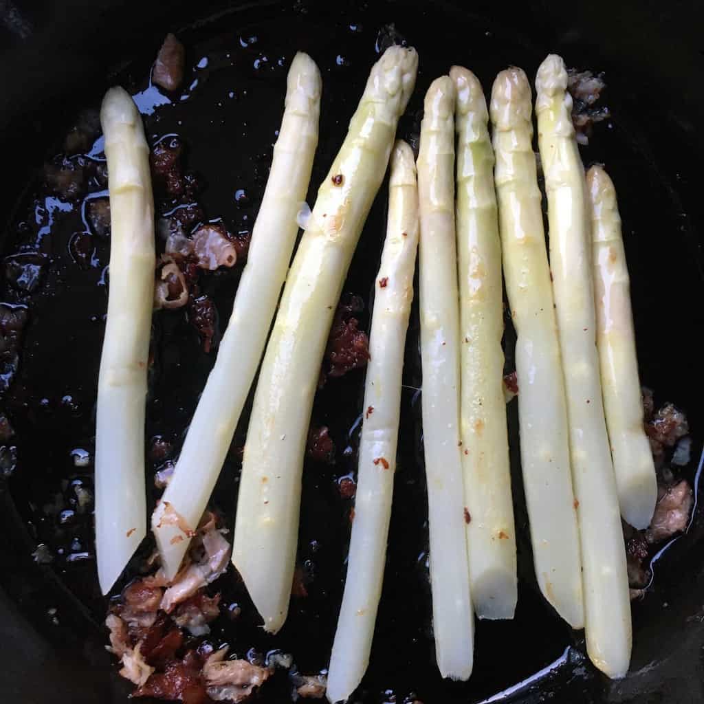 blanched white asparagus in a single layer in a hot cast iron skillet with crispy ham