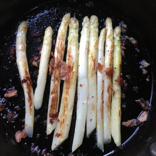 white asparagus cooking in a cast iron skillet with bits of crispy ham sprinkled on top