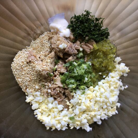 tuna fish salad components in a medium clear glass mixing bowl with ridges pattern with each ingredient in its own small pile one next to the other