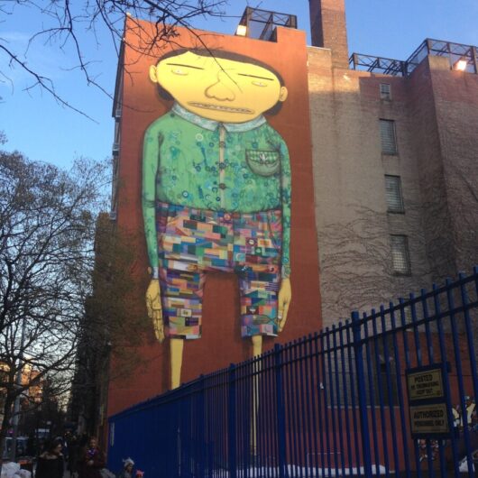 multi-storied building in NYC with an art installation of a school kid with shorts on that are made from a pattern of different flags of the world, a green blouse with pocket and hands at his/her side spanning the height of the entire building
