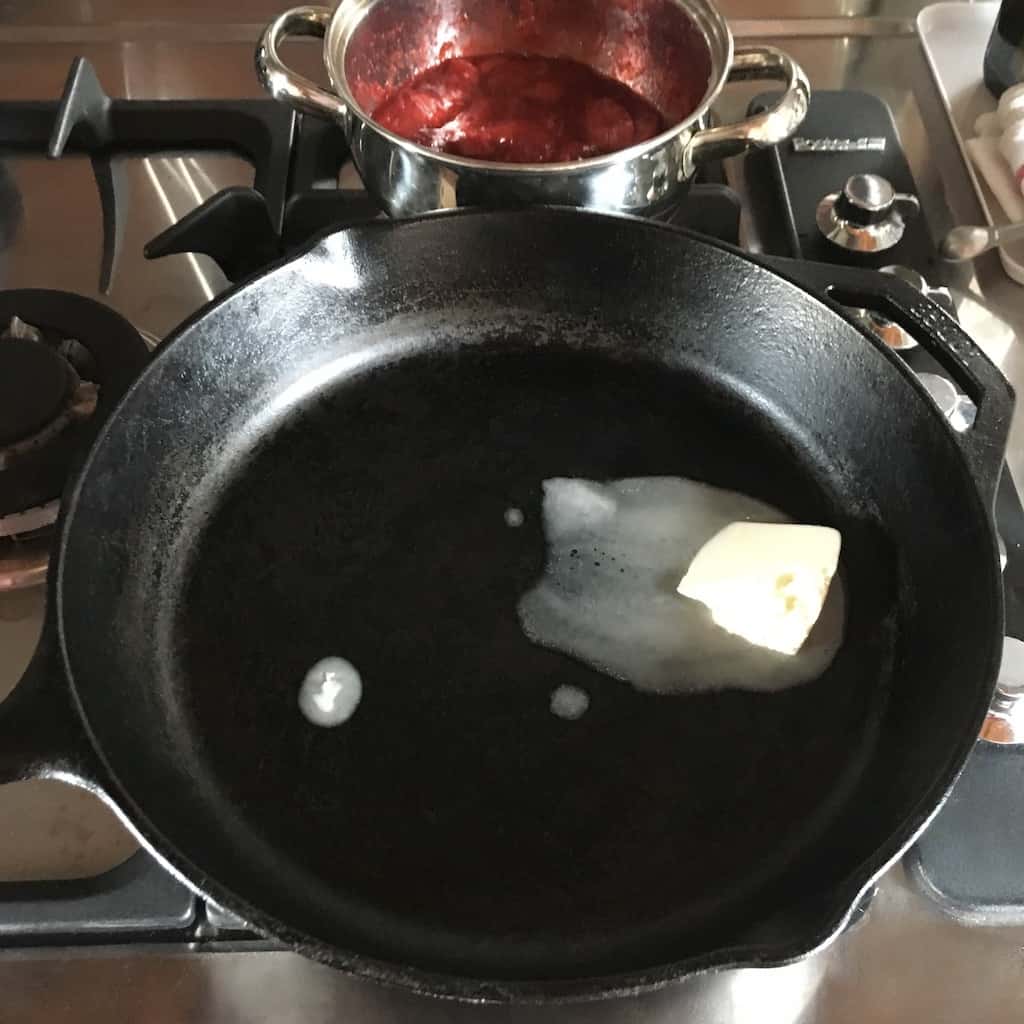 cast iron skillet with a pat of butter melting in it with a pot of strawberry jam sauce in the background