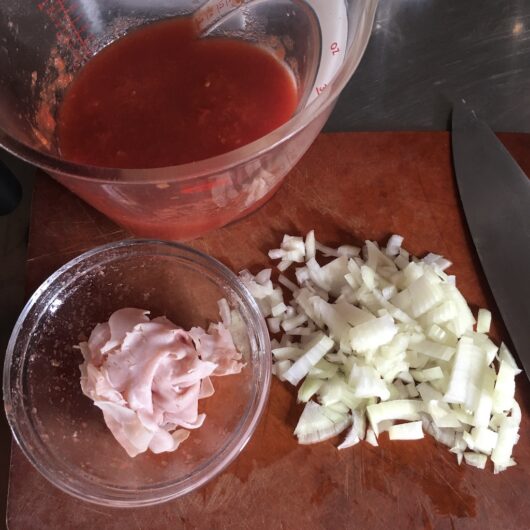 Mutti brand finely chopped tomatoes in an Oxo brand 4 cup measuring cup, a small glass bowl with prosciutto ham in it, and chopped onions all on an Epicurean wood cutting board