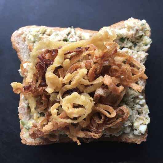 tuna salad spread on one slice of whole wheat bread with crispy french fried onion strings on top