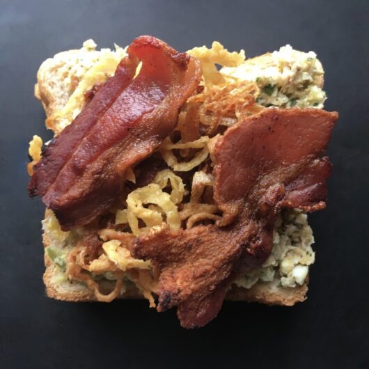 tuna salad spread on one slice of whole wheat bread with crispy french fried onion strings on top with two slices of crispy thick-cut bacon on top