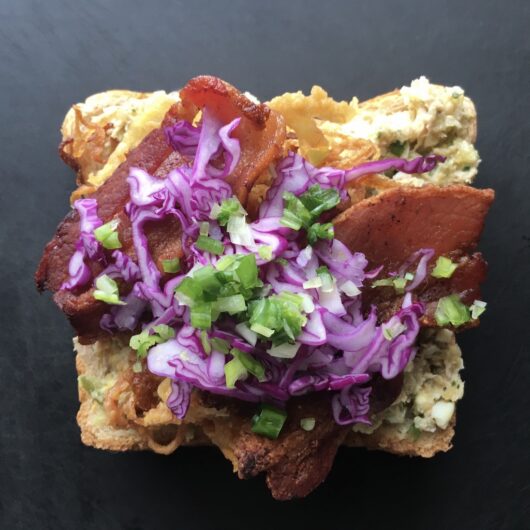 tuna salad spread on one slice of whole wheat bread with crispy french fried onion strings on top with two slices of crispy thick-cut bacon on top and shredded purple cabbage and scallions sprinkled on top