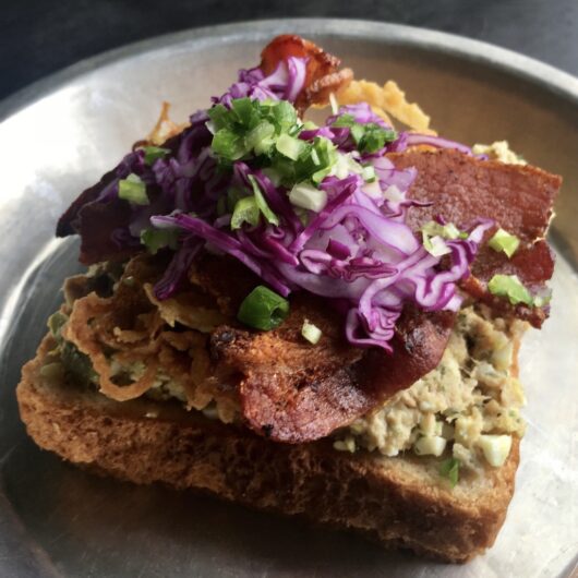 closeup in a vintage pie tin of closeup of tuna salad spread on one slice of whole wheat bread with crispy french fried onion strings on top with two slices of crispy thick-cut bacon on top and shredded purple cabbage and scallions sprinkled on top