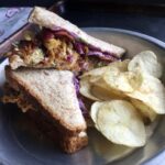 yellowfin tuna sesame scallion salad with crispy bacon, french fried Vidalia onions and purple cabbage strips on wheat bread with a side of plain potato chips in a vintage 9" pie tin with a tea towel with flowers in the background