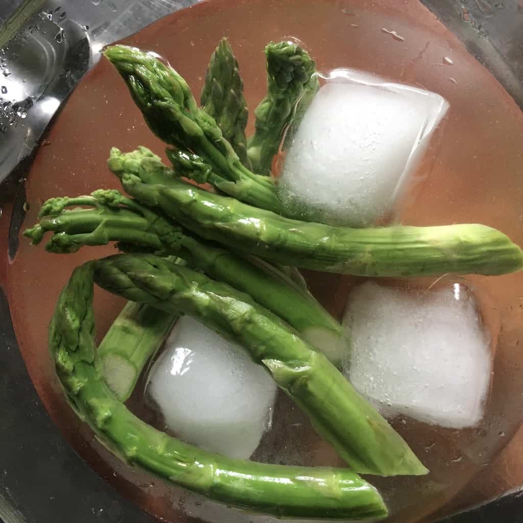 blanched asparagus in bowl of ice water with three king size ice cubes floating in the clear glass bowl