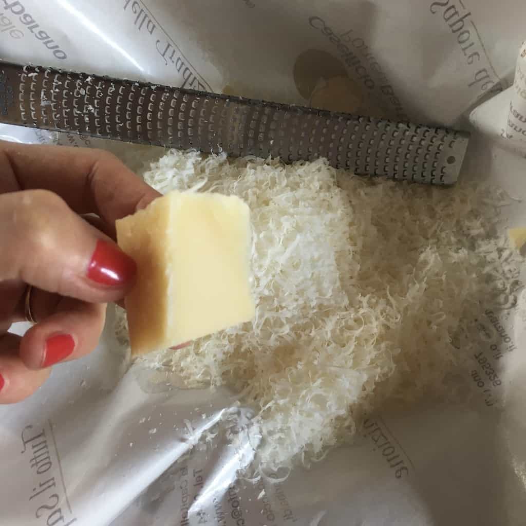 my hand holding a 2 inch piece of hard cheese with a microplane and grated cheese next to it
