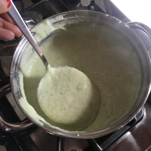 asparagus bechamel in a ladle being held by a hand with red fingernail polish over the pot of bechamel