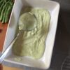a ladle in filled with asparagus bechamel in a white casserole dish with asparagus next to it on a cutting board