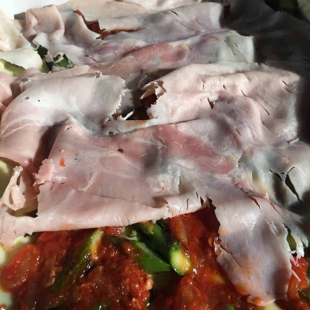 closeup of the prosciutto cotto ham slices being layered over tomato sauce and chopped pan seared asparagus